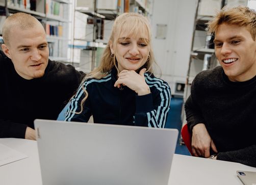 Drei Studierende sitzen vor einem Laptop in der Bibliothek der Deutschen Sporthochschule
