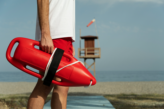 Rettungsschwimmer hält eine rote Rettungsboje am Strand; im Hintergrund stehen Wachturm und Meer.
