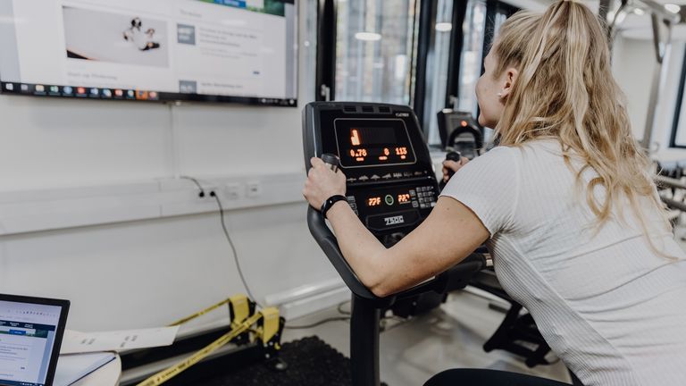 Foto von Frau die in Sportkleidung auf Ergometer sitzt und einen Bildschirm vor ihr betrachtet
