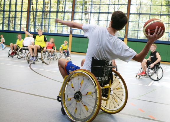 Verschiedene Menschen spielen Rollstuhlbasketball in der Halle. Im Vordergrund ist ein Spieler mit weißem Shirt zu sehen, der gerade zum Wurf ansetzt.
