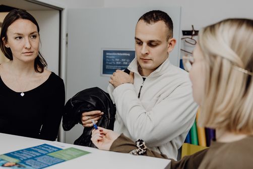 Foto von drei Personen die um einen Tisch mit einem kleinen Flyer sitzen
