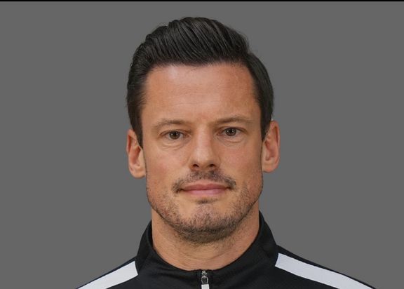Portätfoto von Michael Delura. Er steht vor einem grauen Hintergrund. Er hat eine schwarze Trainingsjacke an mit weißen Streifen auf den Schultern.
