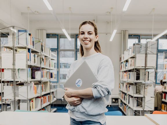 Studentin lächelt mit Laptop in der Hand in der Bibliothek

