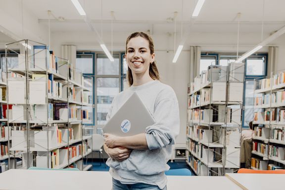 Studentin lächelt mit Laptop in der Hand in der Bibliothek
