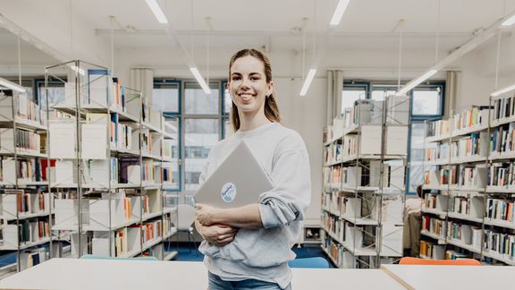 Studentin lächelt mit Laptop in der Hand in der Bibliothek
