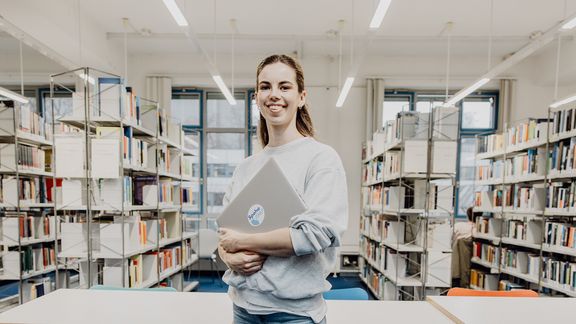 Studentin lächelt mit Laptop in der Hand in der Bibliothek
