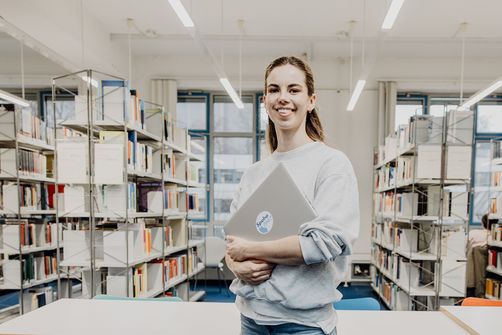 Studentin lächelt mit Laptop in der Hand in der Bibliothek
