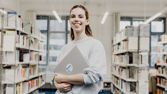 Studentin lächelt mit Laptop in der Hand in der Bibliothek
