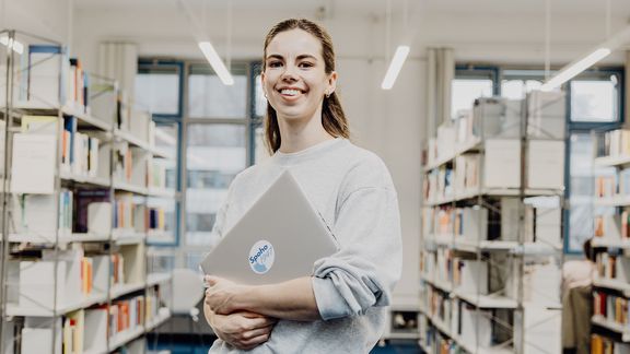 Studentin lächelt mit Laptop in der Hand in der Bibliothek
