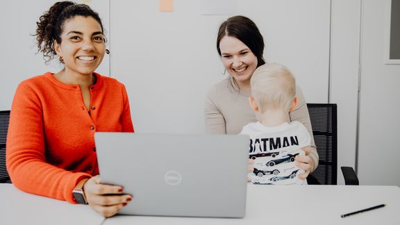 Beratungssituation vor dem Laptop mit der Referentin aus dem Familienservice und einer jungen Frau mit Kind
