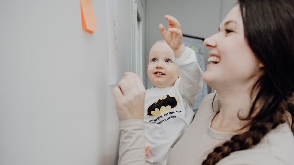 eine junge Frau steht mit Baby auf dem Arm an einem Whiteboard und lacht
