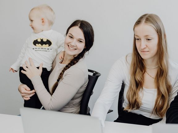 drei junge Menschen sitzen vor einem Laptop gemeinsam an einem Tisch, eine der Frauen hat ein Baby auf dem Arm
