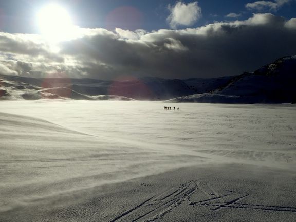 Gruppe von Menschen in weiter, schneebedeckter Landschaft mit starkem Wind und tiefstehender Sonne
