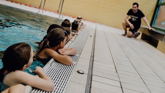 Kinder hören am Beckenrand im Wasser einem Lehrer zu
