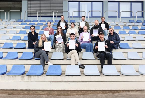 mehrere Personen sitzen auf Besucher*innenrängen eines Stadions und halten Urkunden hoch
