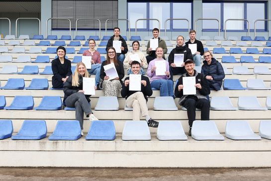 mehrere Personen sitzen auf Besucher*innenrängen eines Stadions und halten Urkunden hoch