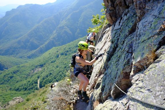 Zwei Personen klettern an einem Fels mit Klettersteigset und Helm in bergiger Landschaft
