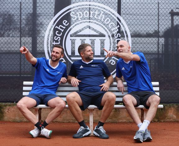 Drei Männer in Sportkleidung sitzen auf einer weißen Bank auf einem Tennisplatz und zeigen in verschiedene Richtungen. Im Hintergrund ist das Logo der Sporthochschule zu sehen. 
