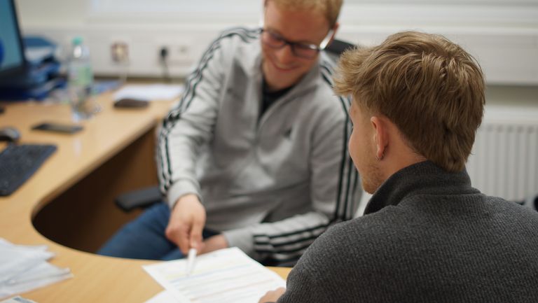 Hier wird eine Beratungsszene gezeigt mit zwei sehr gut Aussehenden Mitarbeitern des Zentrums für Sportleher*innenbildung
