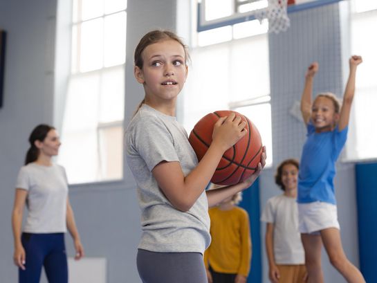 Schülerin mit Basketball in den Händen schaut zurück; Lehrerin und weitere Schüler im Hintergrund
