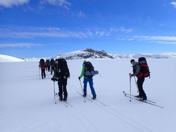 Gruppe von Skifahrern mit Rucksäcken quert verschneite, weite Landschaft unter blauem Himmel
