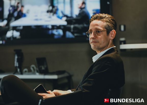 Porträtfoto von Philipp Havermann. Er sitzt mit einem schwarzen Jackett und seinem Handy in der Hand in einem Raum. Im Hintergrund sind ein Laptop und ein großer Fernseher, sowie eine Lampe zu sehen.
