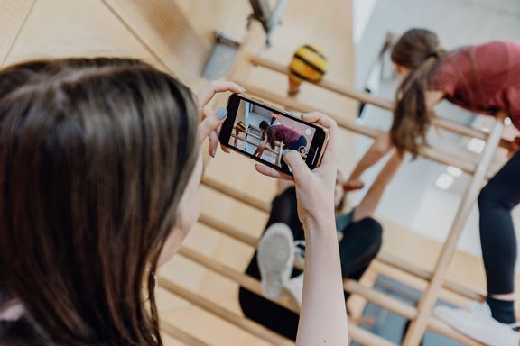 Eine Person filmt mit einem Smartphone Kinder beim Klettern an einer Sprossenwand in einer Turnhalle. Auf dem Display ist die Aufnahme der Bewegungssituation zu sehen.
