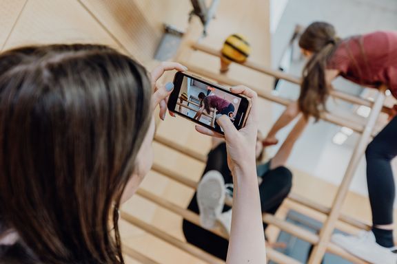 Eine Person filmt mit einem Smartphone Kinder beim Klettern an einer Sprossenwand in einer Turnhalle. Auf dem Display ist die Aufnahme der Bewegungssituation zu sehen.
