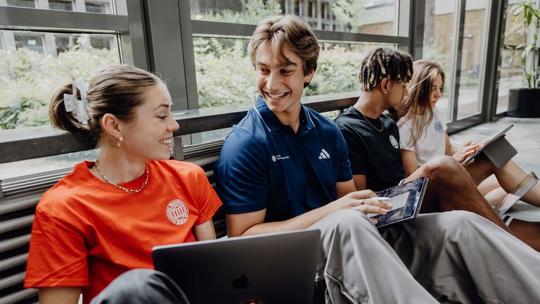 Vier Studierende sitzen auf dem Boden des Foyers und arbeiten mit Laptops und Tablets.
