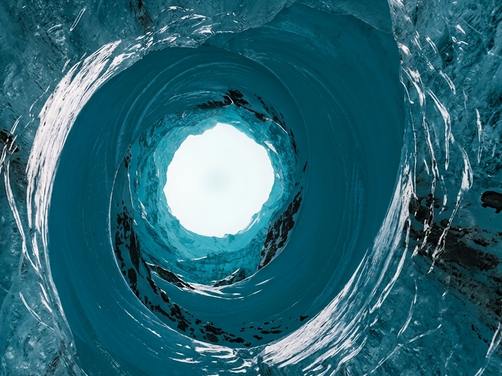 Blick von unten durch ein rundes Loch in einer Eisschicht auf den Himmel
