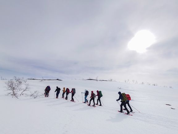 Gruppe von Menschen mit Schneeschuhen wandert in einer verschneiten Berglandschaft bei bewölktem Himmel
