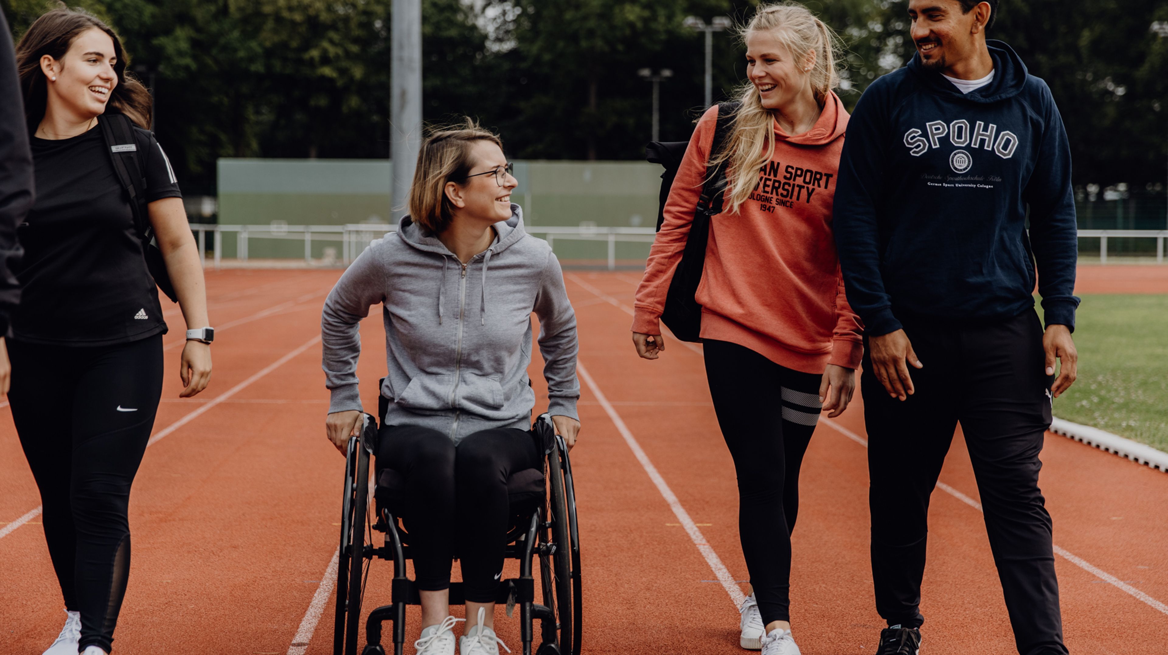 Studierende im Rollstuhl fährt neben laufenden Studierenden auf der Laufbahn der Deutschen Sporthochschule
