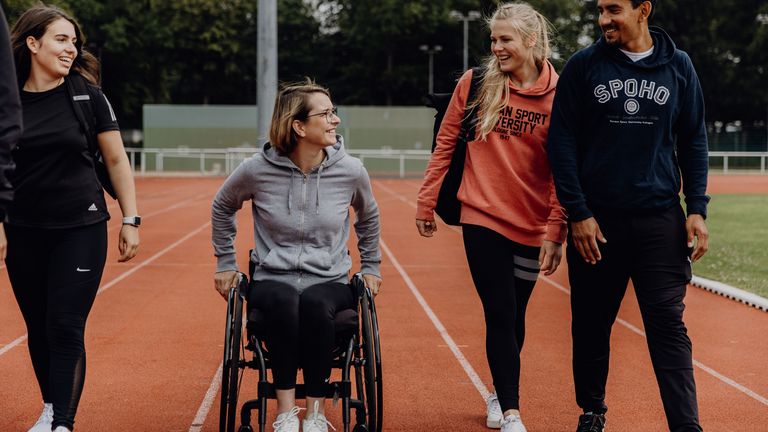 Studierende im Rollstuhl fährt neben laufenden Studierenden auf der Laufbahn der Deutschen Sporthochschule
