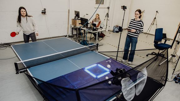 Probandin führt Tischtennisaufschlag auf einer Platte in Richtung eines leuchtenden Quadrats auf der anderen Seite aus
