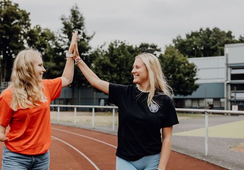 Zwei Studentinnen geben sich auf einer Laufbahn im Freien ein High-Five.
