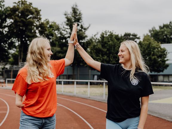 Zwei Studentinnen geben sich auf einer Laufbahn im Freien ein High-Five.
