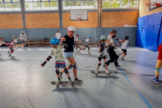 Eine Gruppe von Kindern lernt Skateboardfahren in einer Schulsporthalle.