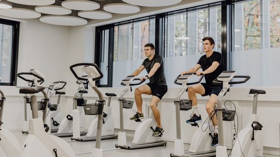 Foto von zwei Männern die auf Ergometern in einem Raum mit vielen weiteren Ergometern sitzen
