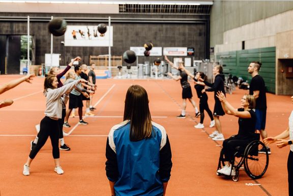 Lehre in der Halle, die Dozentin steht im Vordergrund von hinten sichtbar, die Studierende stehen sich paarweise gegenüber und werfen sich einen Ball zu. Eine Studierende sitzt im Rollstuhl.
