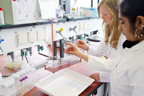 Zwei Schülerinnen im weißen Kittel stehen in einem Labor und arbeiten mit einer Pipette
