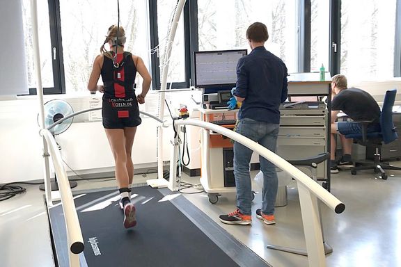 Man sieht eine Läuferin bei einer spiroergmetrischen Laufbanduntersuchung. Man sieht sie die Läuferin von hinten, sie trägt eine Spiromaske und ist verkabelt. Neben dem Laufband steht eine Person, die an einem Bildschirm die Werte überwacht.
