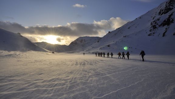 Eine Gruppe Studierende wandert im Schnee dem Sonnenuntergang entgegen. Links und rechts sind verschneite Berge zu sehen.
