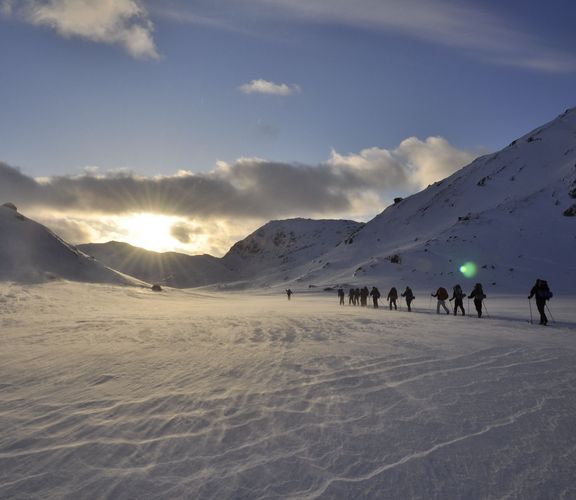 Eine Gruppe Studierende wandert im Schnee dem Sonnenuntergang entgegen. Links und rechts sind verschneite Berge zu sehen.

