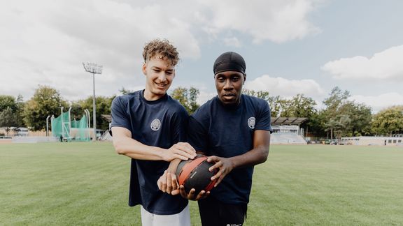 Foto von zwei Männern die auf einem Rasenplatz sich einen Ball Rücken an Rücken seitlich übergeben
