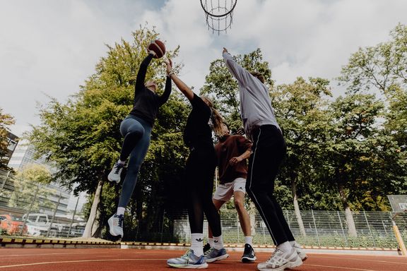 Foto von Basektballspielszene bei der eine Person mit Ball in der Hand hochspringt und von weiteren Personen verteidigt wird
