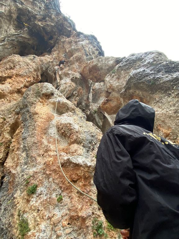 Blick von unten auf eine Felswand mit einem Kletterer im Überhang und einer Person in schwarzer Jacke im Vordergrund
