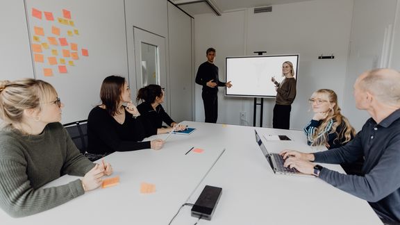 Zwei Personen präsentieren in einem vollen Besprechungsraum an einem Bildschirm
