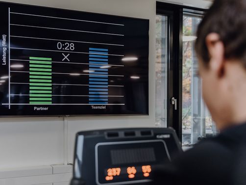 Foto von Person die auf Ergometer in Richtung eines Bildschirms auf dem ein Leistungsdiagramm angezeigt wird sitzt
