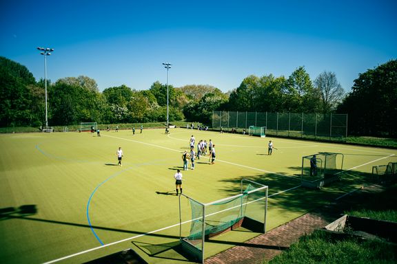 Zu sehen ist ein grüner Hockeyplatz aus der Vogelperspektive, auf dem viele Menschen Hockey spielen.  
