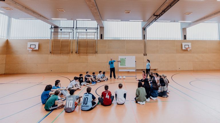 Lehrer zeigt vor Schülern und Schülerinnen auf Plaktat in einer Sporthalle  
